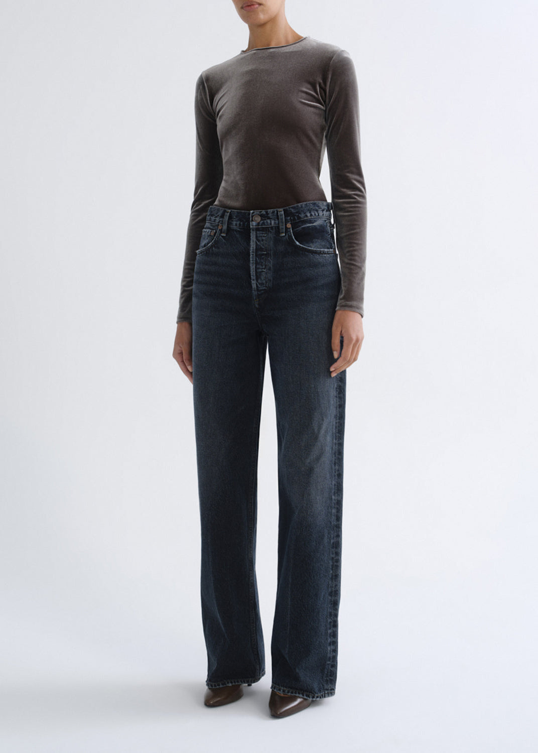 A model wearing a dark brown long-sleeve knit top and dark blue wide-leg jeans, standing against a light gray studio background.