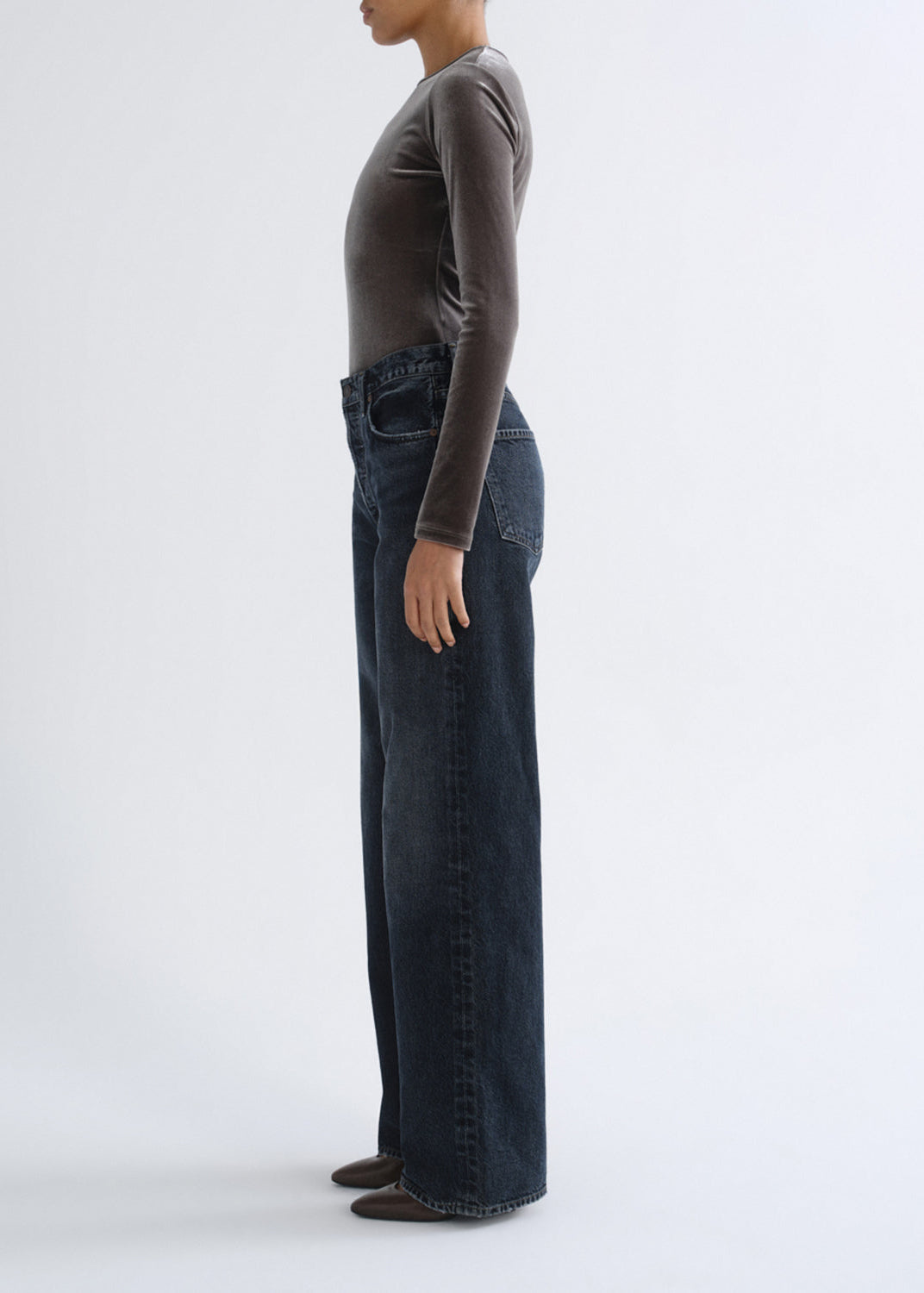 A model shown in profile wearing dark blue wide-leg denim jeans and a dark brown long-sleeve top against a plain white studio background.