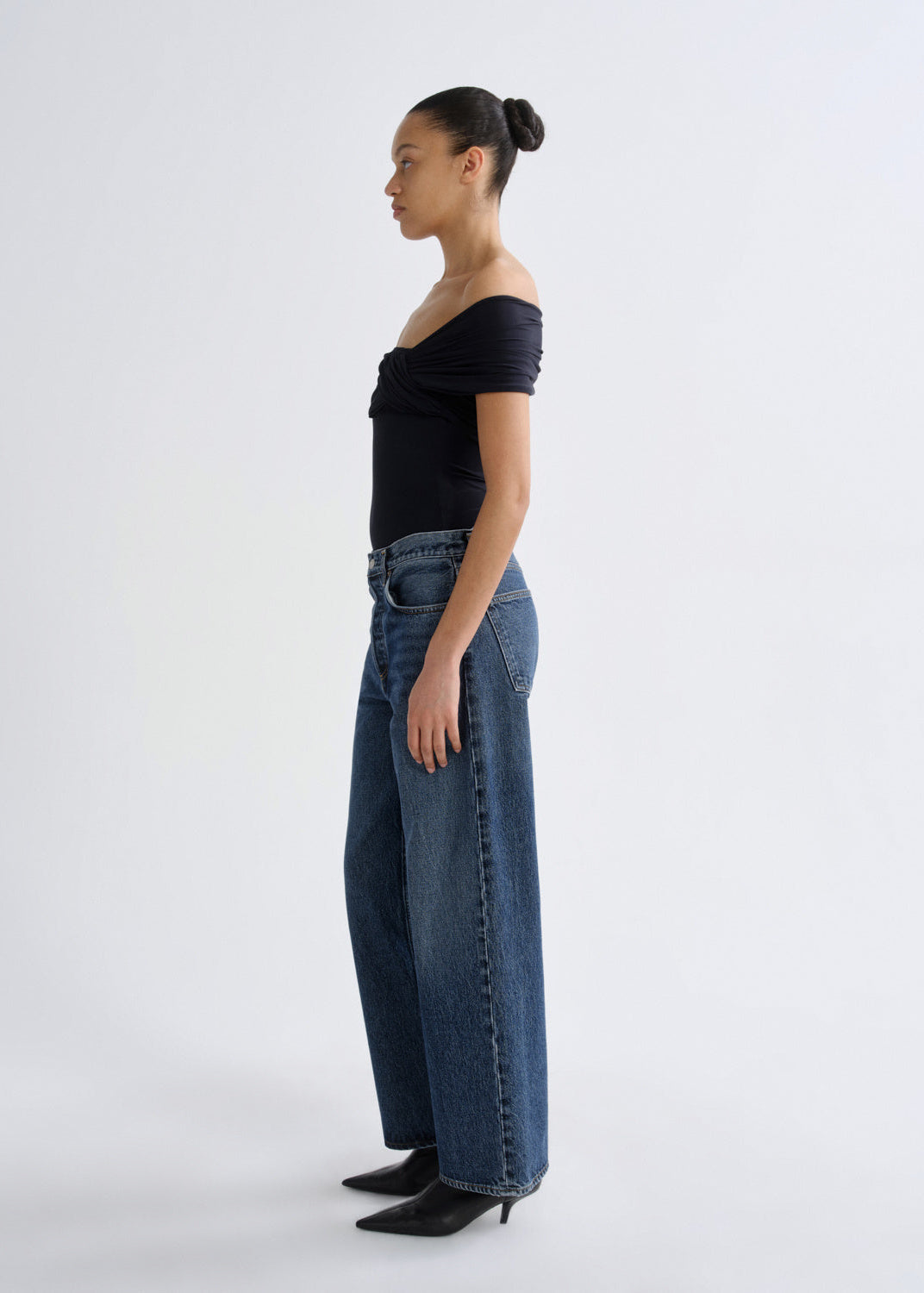 A model shown in profile wearing a black off-the-shoulder short-sleeve top, high-waisted wide-leg blue jeans, and black pointed-toe heels, standing against a plain light gray studio background.