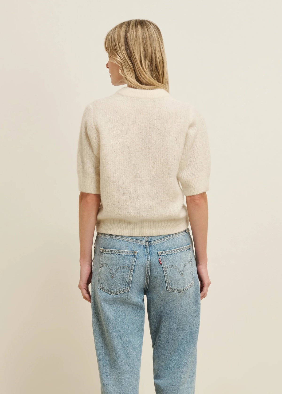 A woman viewed from behind wearing a cream-colored short-sleeve knit sweater and light-wash blue jeans against a plain beige studio background.
