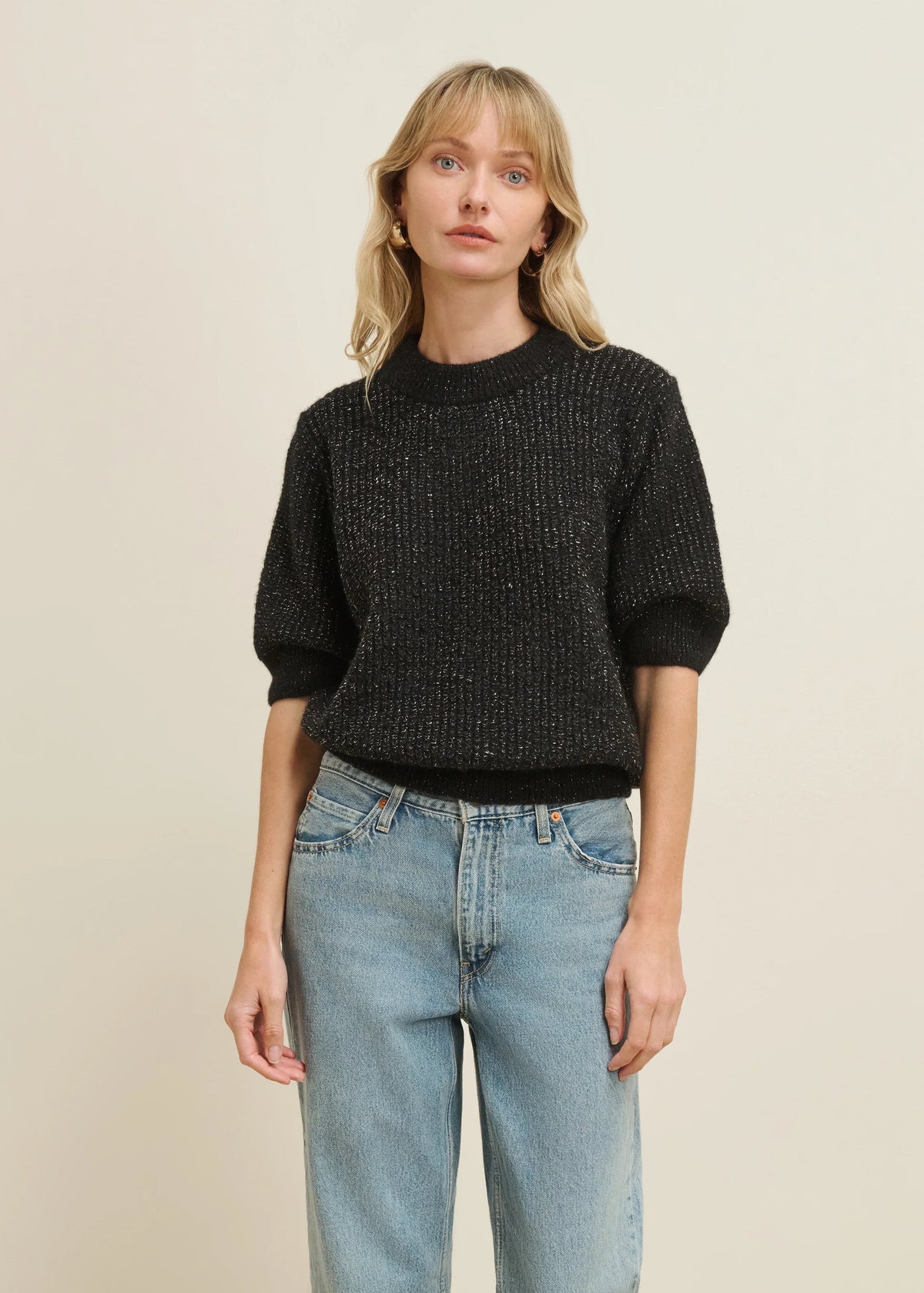 A woman modeling a short-sleeve black knit sweater with a textured, sparkly finish, paired with light-wash blue jeans, against a plain beige background.