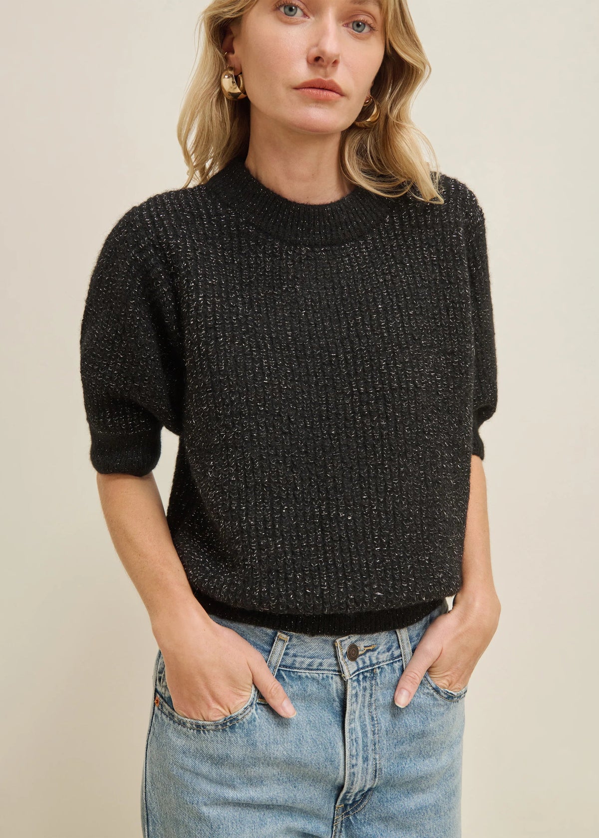 A woman wearing a black textured knit short-sleeve sweater and light blue jeans, standing against a plain beige background.