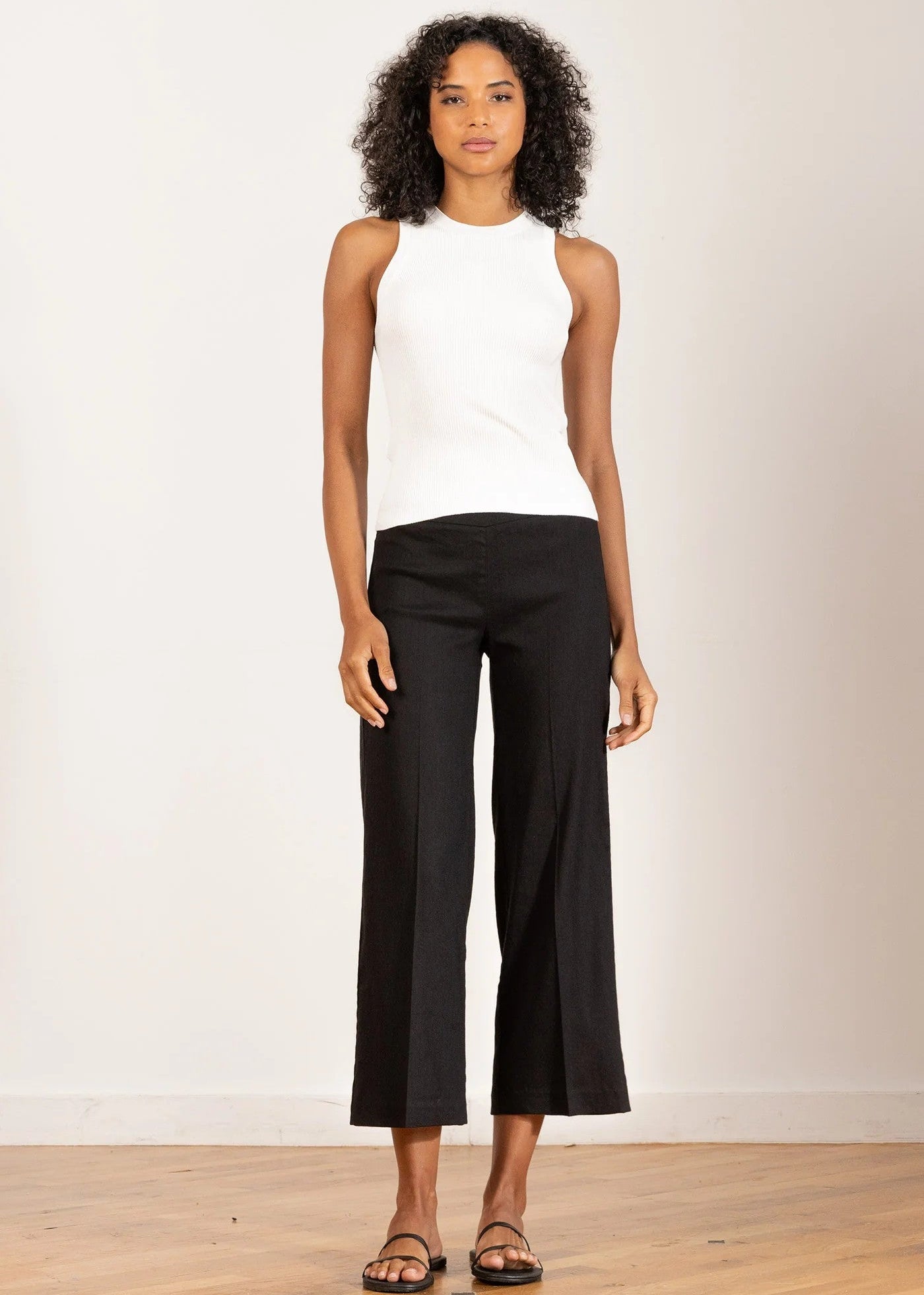 Woman wearing a white sleeveless top and black pants on a plain background