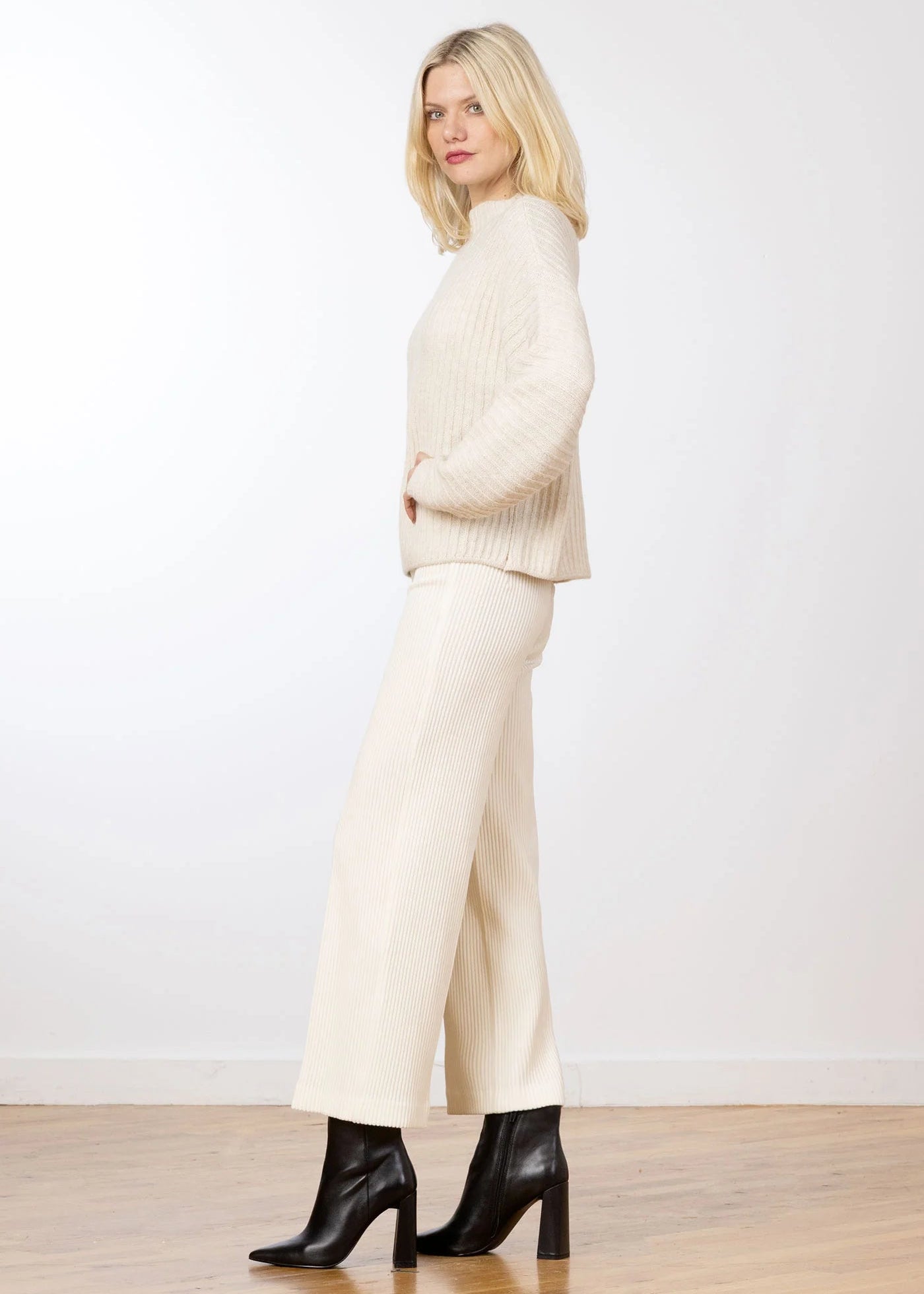 A blonde woman stands in profile wearing a cream ribbed knit mock-neck sweater, matching cream wide-leg trousers, and black heeled ankle boots against a white studio wall and light wood floor.