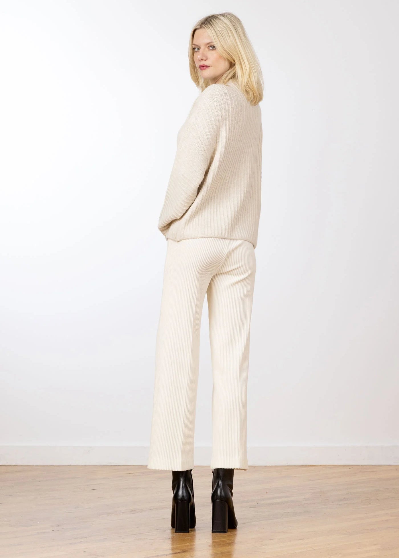 A blonde woman is shown from the back wearing a cream-colored ribbed knit sweater and matching wide-leg trousers, paired with black high-heeled boots, set against a plain white wall and light wood floor.