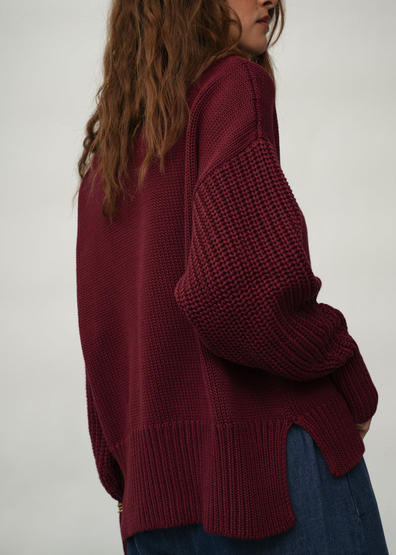A woman wearing a chunky knit maroon sweater and blue jeans, shown from a side-back angle against a plain light gray background.