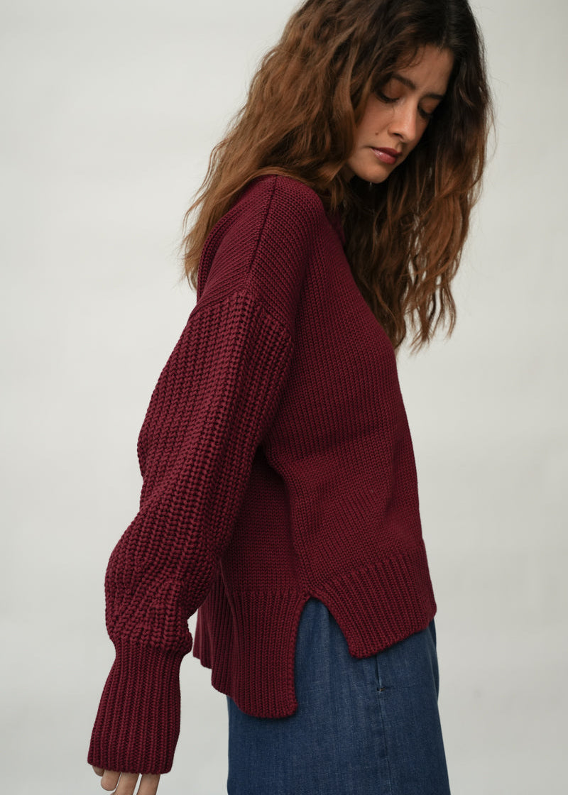 A woman wearing a chunky knit sweater in deep burgundy, paired with blue jeans, photographed against a neutral light gray background.