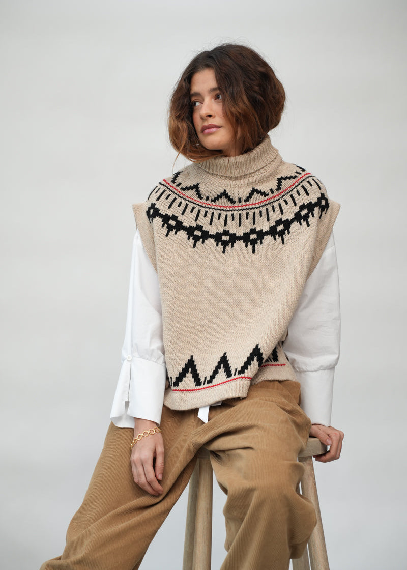 A woman seated on a stool wearing a beige knit turtleneck sweater vest with a black and red geometric pattern, layered over a white long-sleeve top and tan pants, against a plain light gray studio background.