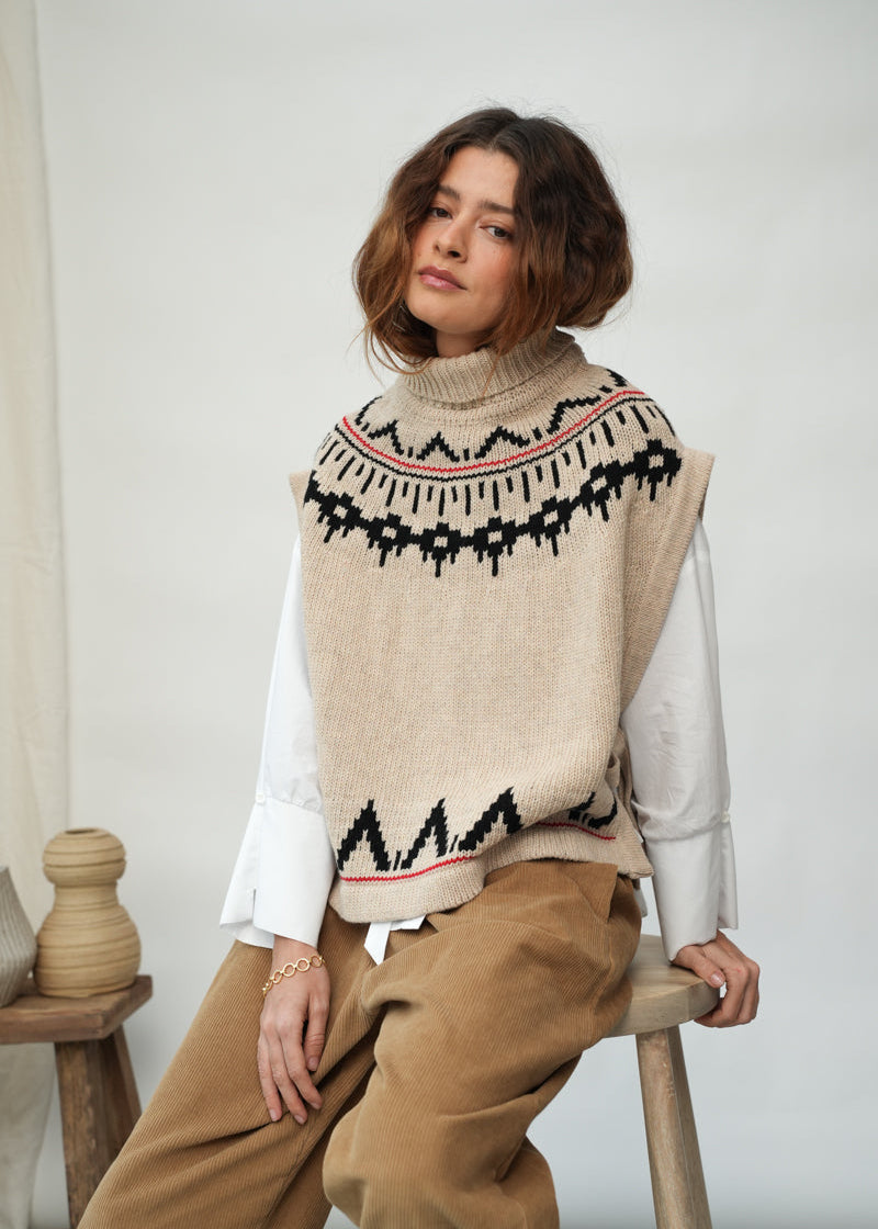A woman seated on a wooden stool wearing a beige knit sweater vest with a black and red geometric pattern over a white long-sleeve top and tan pants, photographed in a softly lit indoor setting.