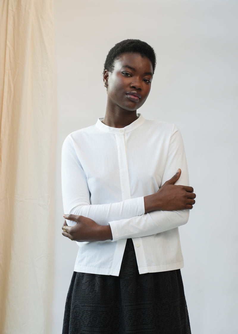 A model wearing a white long-sleeve button-up shirt and a black textured skirt, standing with arms crossed against a neutral studio backdrop.