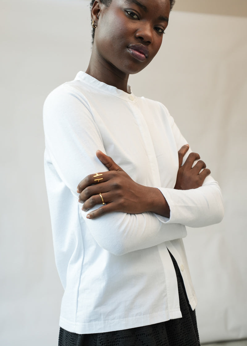 A model wearing a white long-sleeve mock-neck top with a side slit, paired with a black skirt, standing with arms crossed against a plain light background.