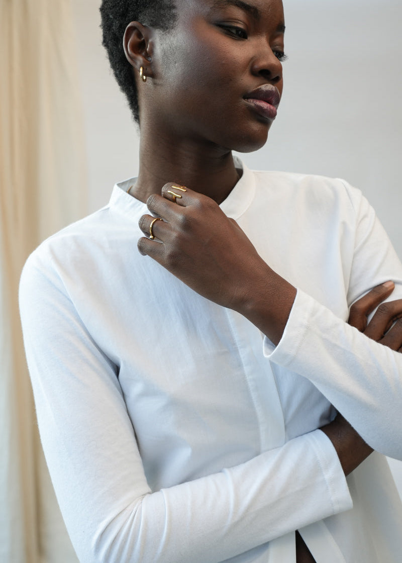 A woman wearing a white long-sleeve button-up shirt, shown from the chest up with her arms crossed and one hand near her collarbone. She is wearing gold rings and a small gold hoop earring.