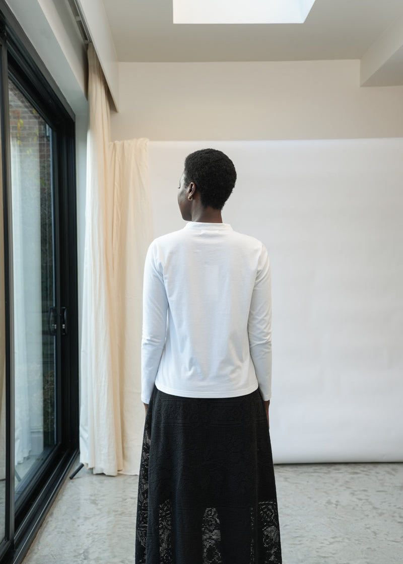 A person viewed from behind wearing a white long-sleeve mock-neck top and a long black skirt with lace detailing, standing in a minimalist room with a white backdrop, window, and curtains.