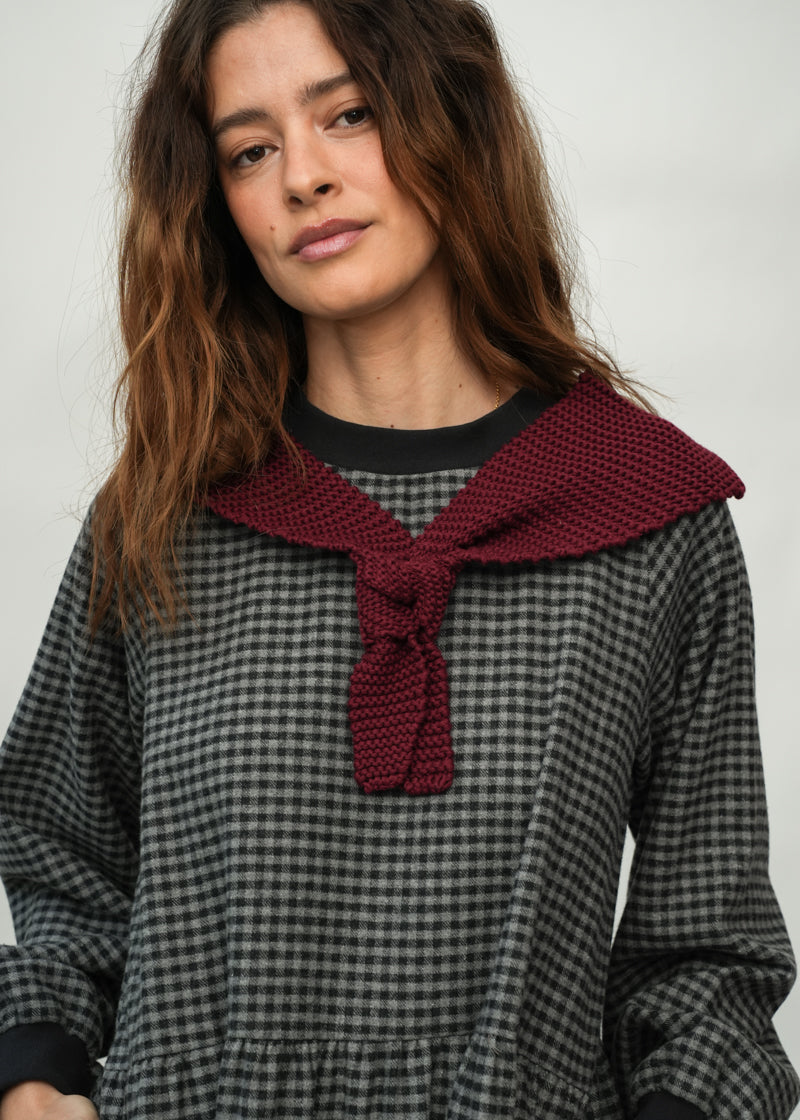 A woman wearing a black and gray gingham long-sleeve dress with a large burgundy knit collar tied in a knot at the neck, shown against a plain light gray background.