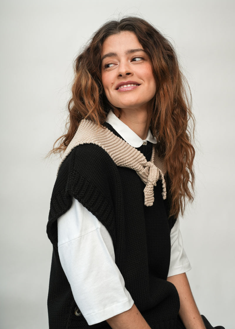 A woman with wavy brown hair wearing a black knit vest over a white collared shirt, with a beige knit sweater draped around her shoulders, photographed against a plain light gray background.