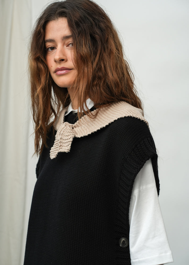 A woman with long, wavy brown hair is shown from the chest up, wearing a black sleeveless knit vest over a white short-sleeve shirt. The vest features a wide, textured beige collar with a decorative bow detail at the center.