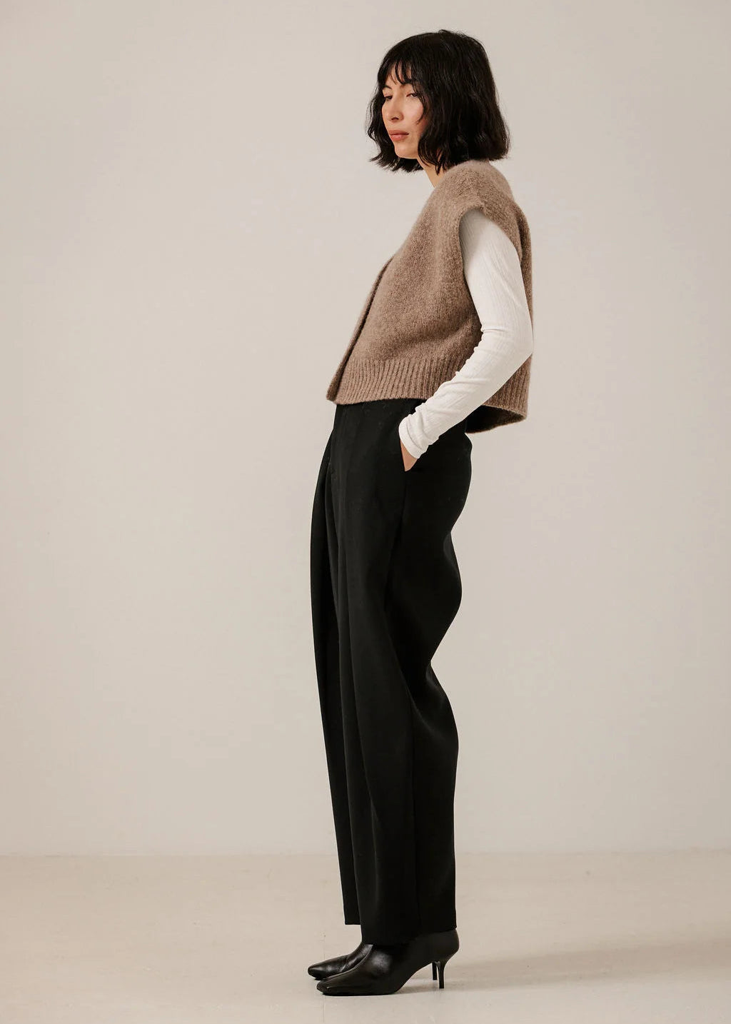 A woman stands in profile wearing a sleeveless taupe knit vest over a white long-sleeve top, black wide-leg trousers, and black pointed heels against a plain light beige studio background.