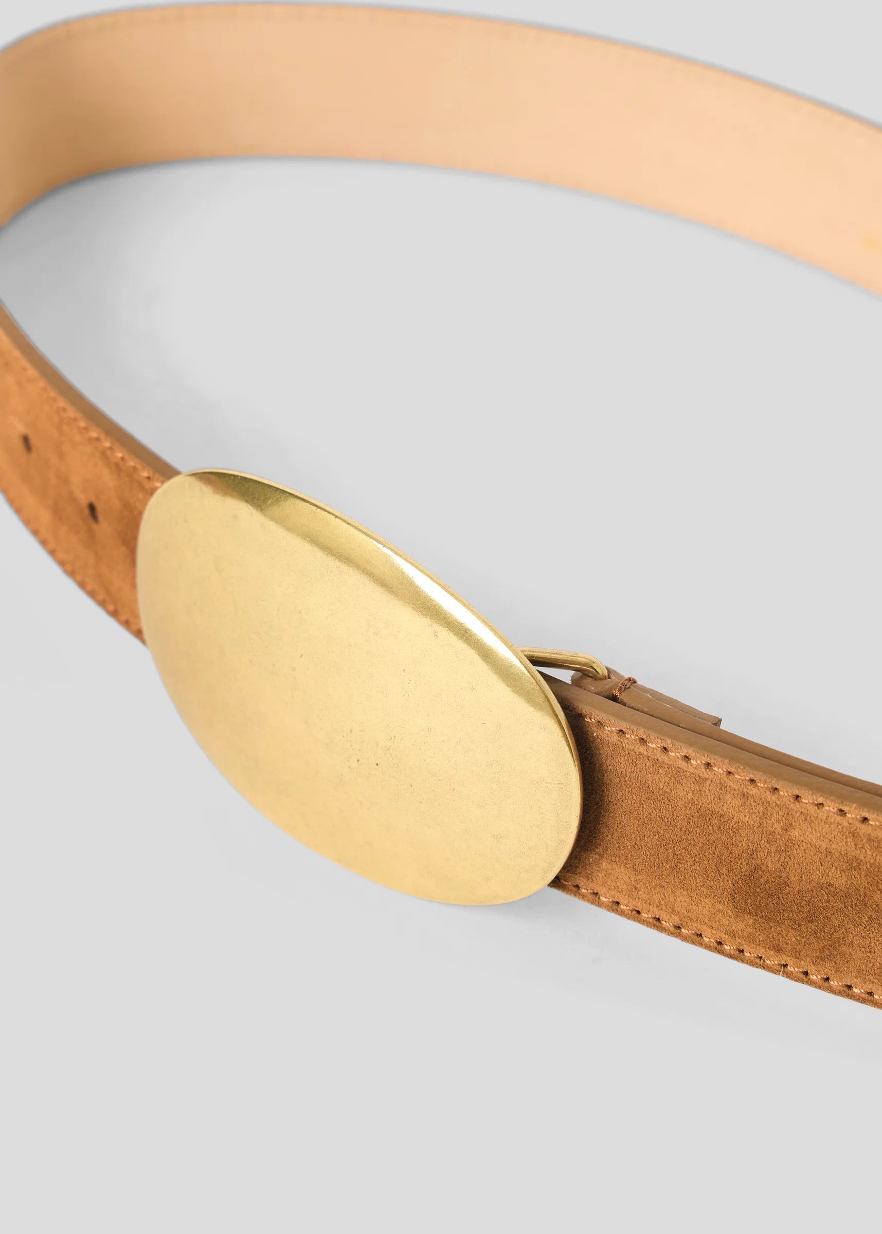 A close-up of a tan leather belt with a large, polished oval gold buckle, set against a neutral light gray background.