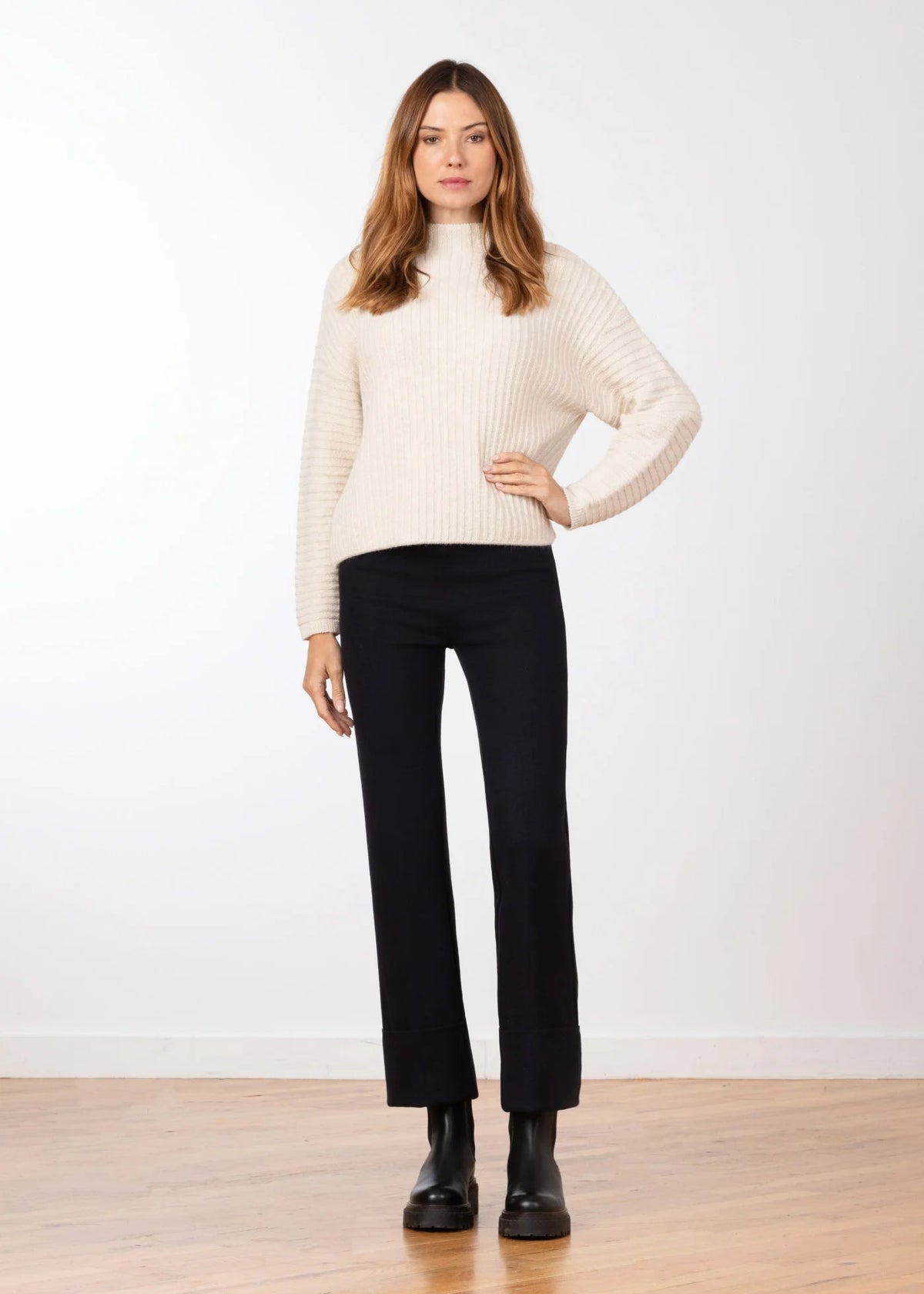 A woman stands in a studio wearing a cream ribbed turtleneck sweater, black cropped trousers, and black ankle boots against a white wall and light wood floor.