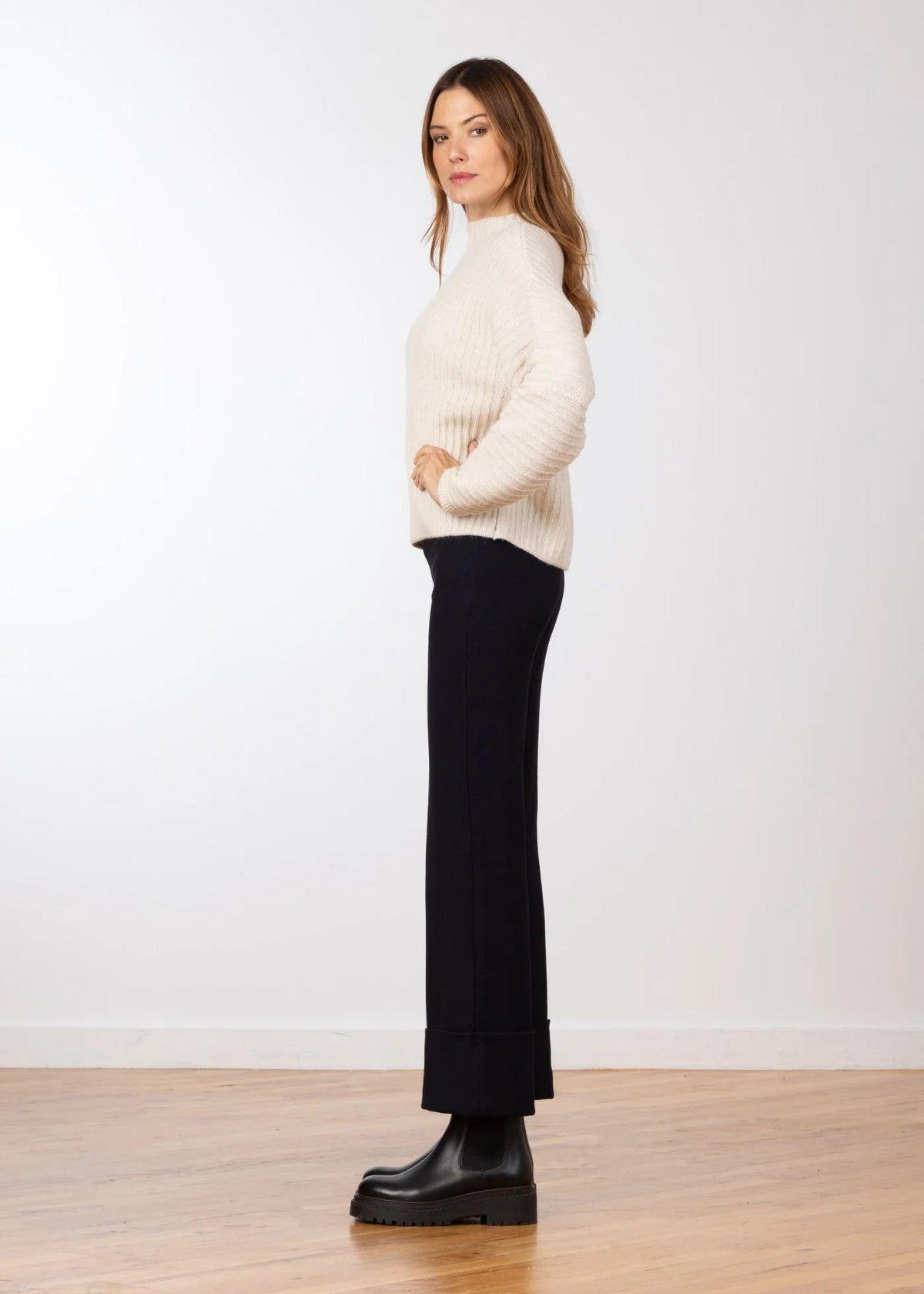 A woman stands in profile wearing a cream ribbed turtleneck sweater, black wide-leg cropped pants, and black platform boots against a white studio wall and light wood floor.