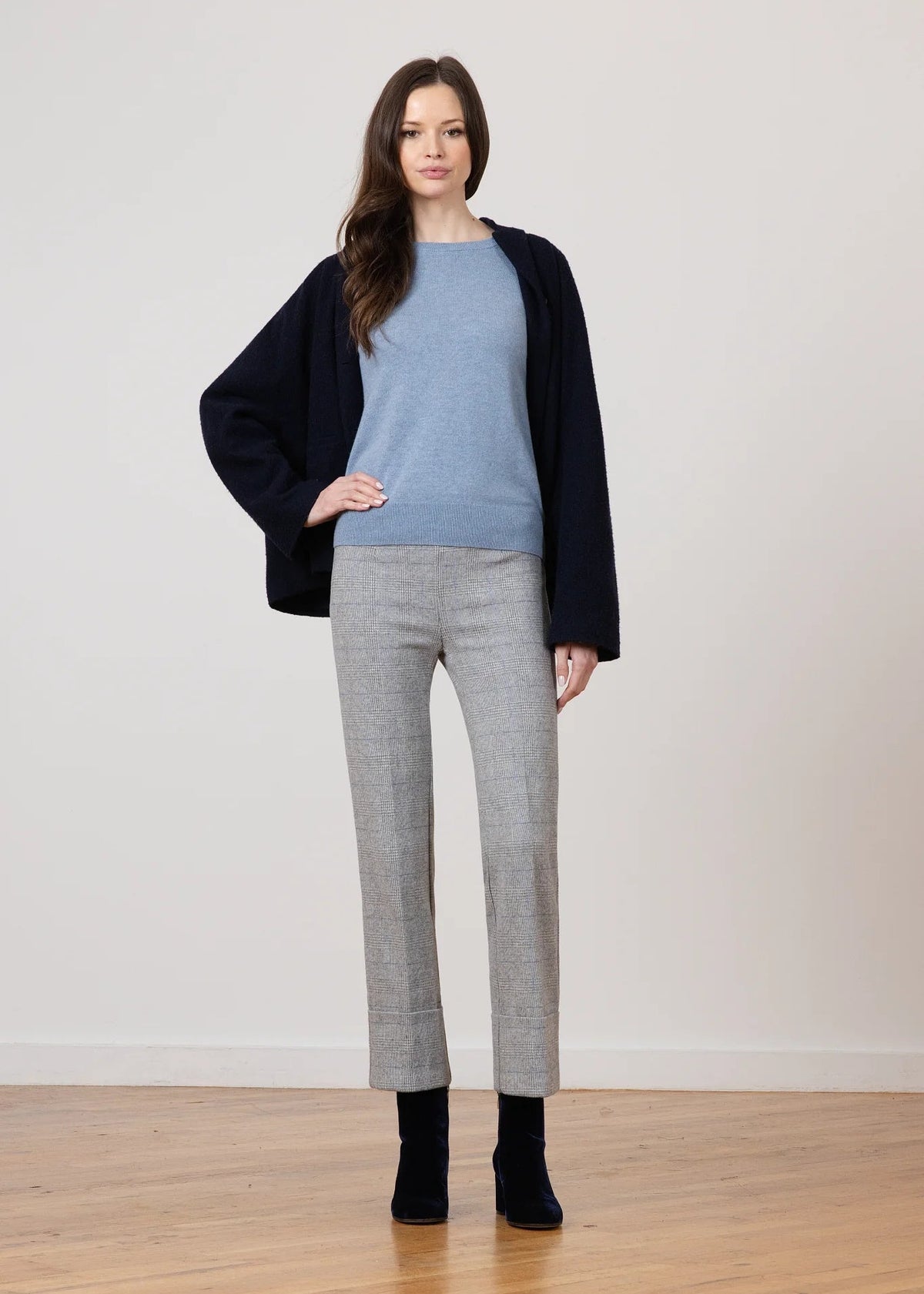 A woman stands in a studio wearing a light blue knit top, a dark navy cardigan, light gray cropped trousers, and black ankle boots against a plain off-white wall and wood floor.