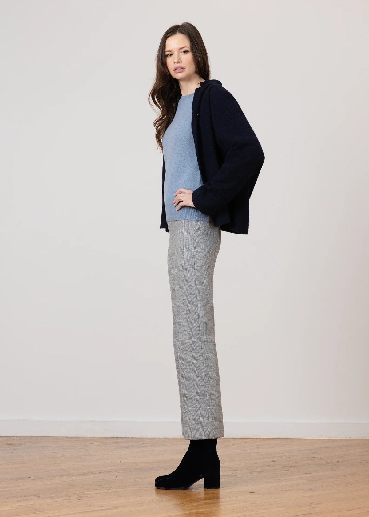 A woman stands in profile wearing a light blue knit sweater, a dark navy cardigan, a light gray textured midi skirt, and black ankle boots, set against a neutral off-white wall and light wood floor.