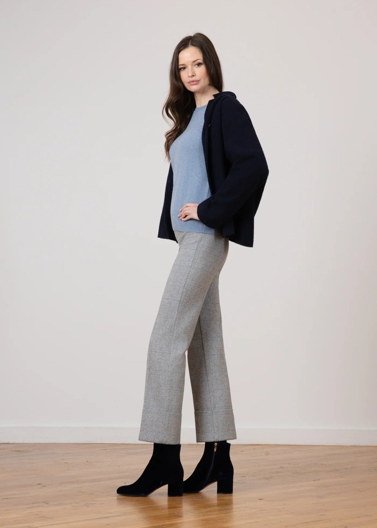 A woman models a stylish outfit consisting of a light blue knit sweater, a dark navy cardigan, wide-leg gray trousers, and black ankle boots, standing against a neutral off-white wall and wooden floor.