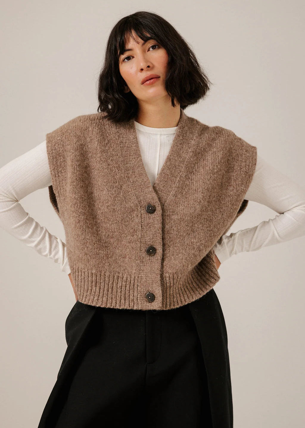 A woman modeling a sleeveless taupe knit vest with a button-front closure over a white long-sleeve top and black skirt, against a plain light gray studio background.