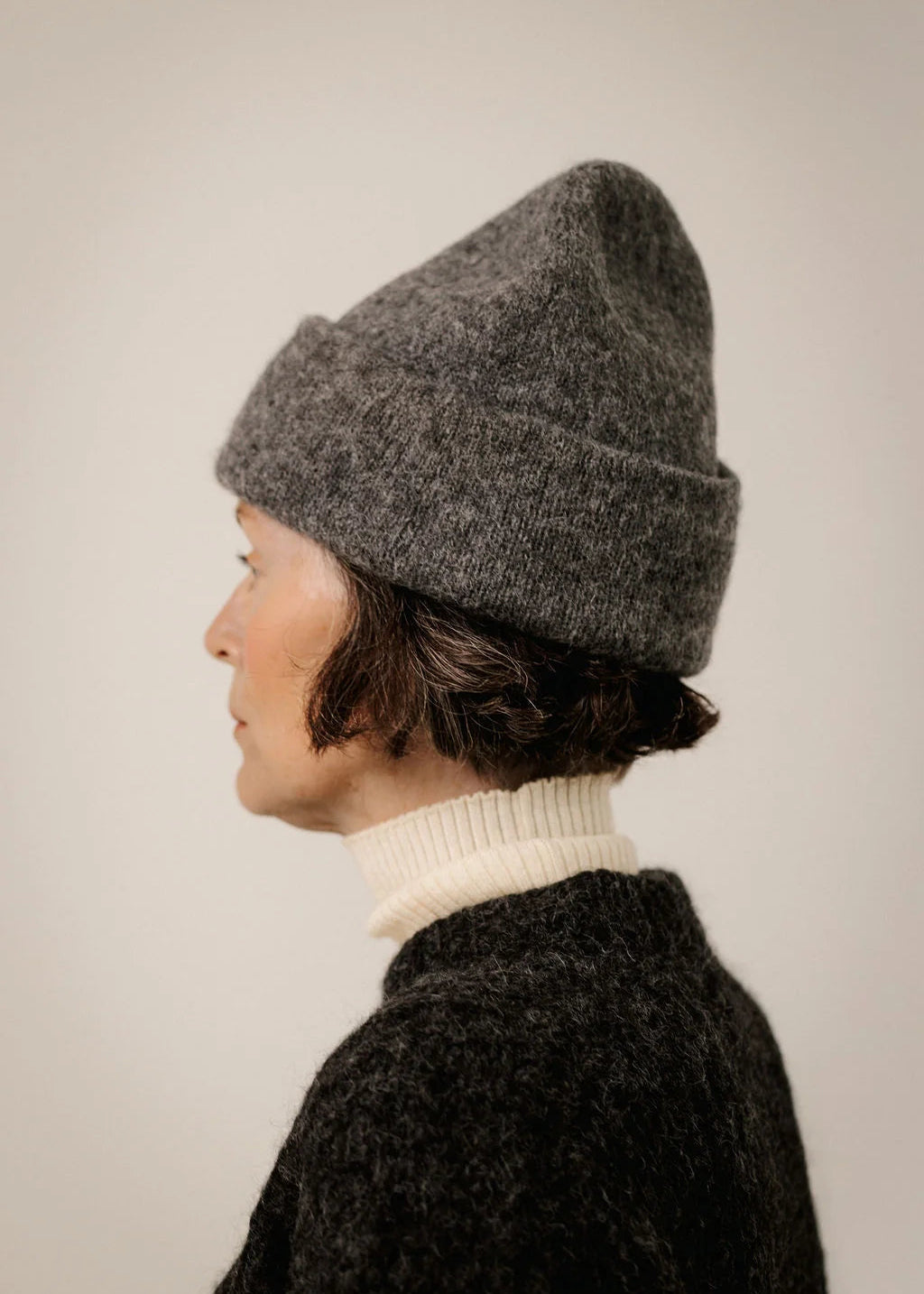 A person shown in side profile wearing a dark heather gray knit beanie, a cream ribbed turtleneck, and a dark textured sweater against a plain light beige background.