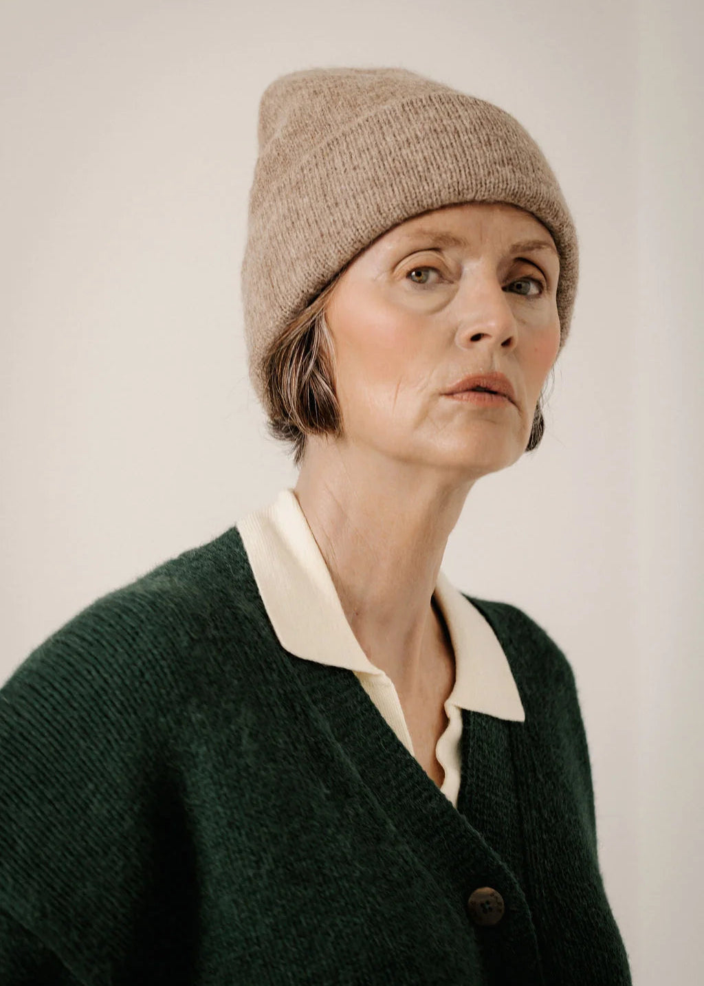 A woman wearing a beige knit beanie and a dark green cardigan over a cream collared shirt, photographed against a plain light background.