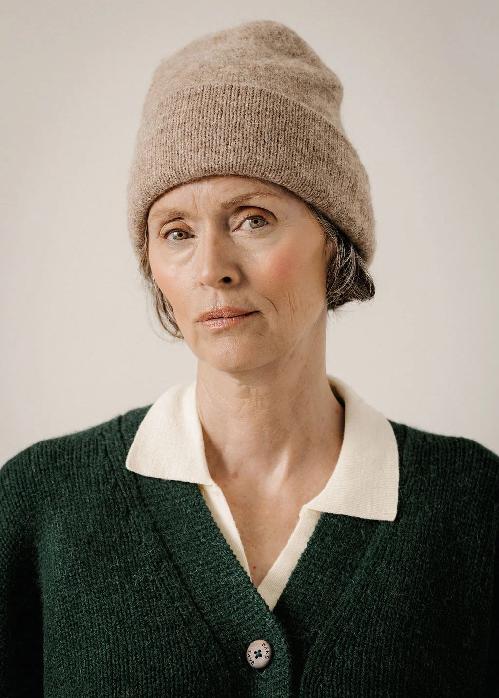 A woman wearing a beige knit beanie and a dark green cardigan with a cream collar, photographed against a plain light background.