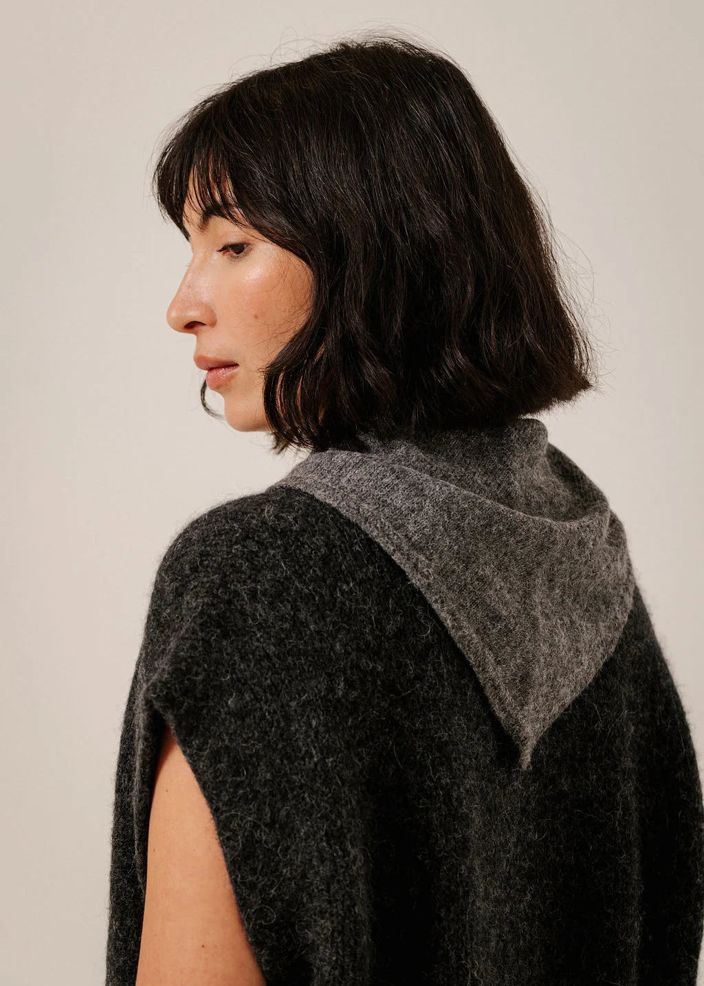 A woman shown from the side and back wearing a dark charcoal gray sleeveless knit top with a contrasting medium gray hood, set against a plain light beige background.