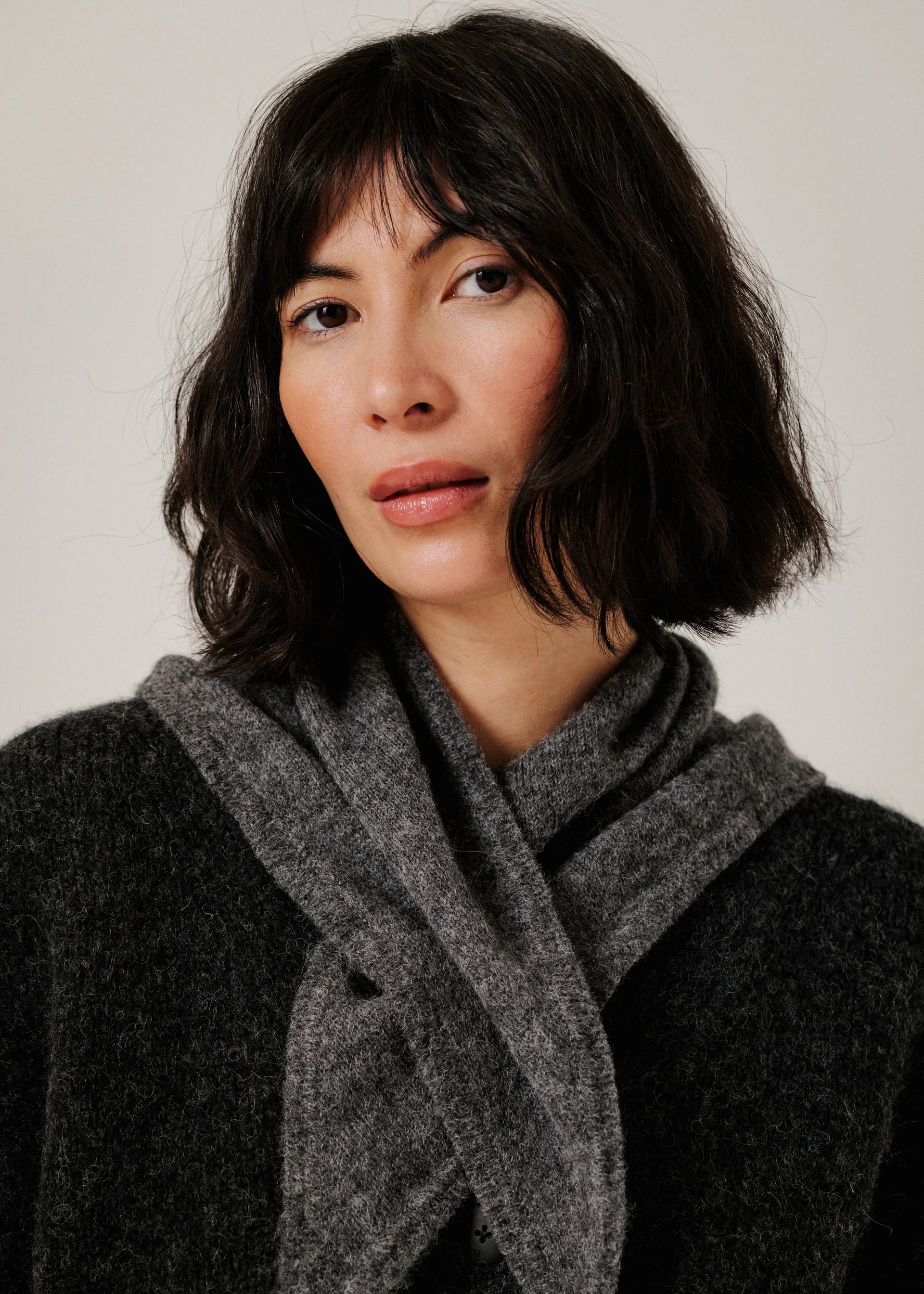 A woman with dark shoulder-length hair wearing a dark charcoal knit sweater and a thick, textured gray knit scarf wrapped around her neck, photographed against a plain light gray background.