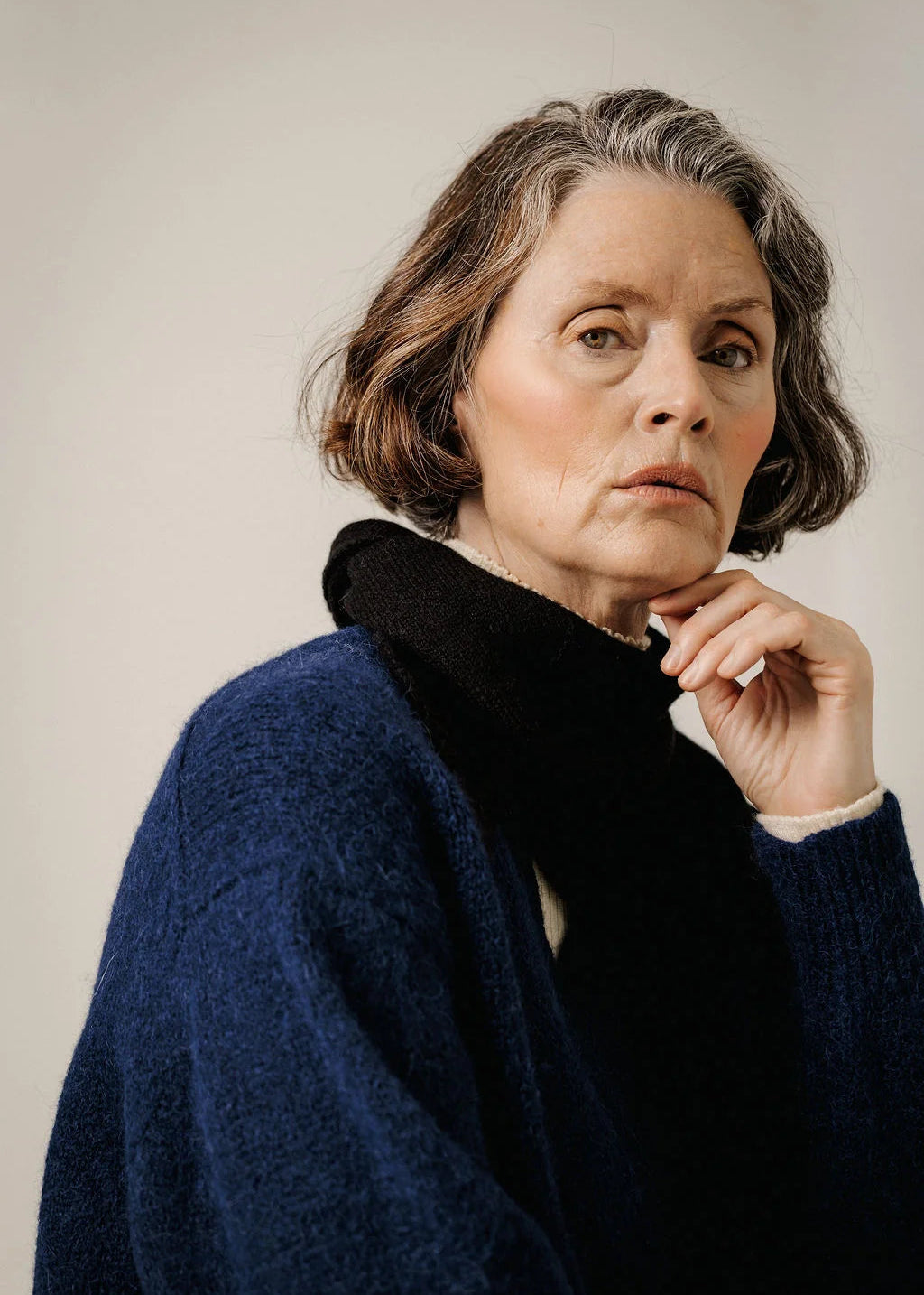 A close-up portrait of a woman with short gray-brown hair wearing a textured navy blue knit sweater and a black fuzzy collar, posed against a plain light beige studio background.