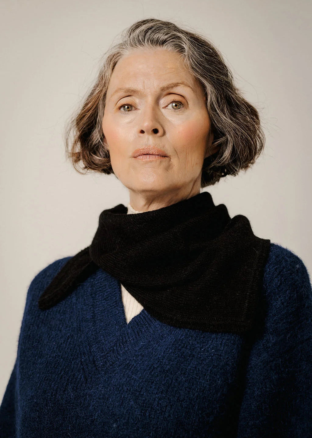 An older woman with short gray hair wearing a black knit cowl scarf and a textured navy blue sweater, photographed against a plain light gray studio background.