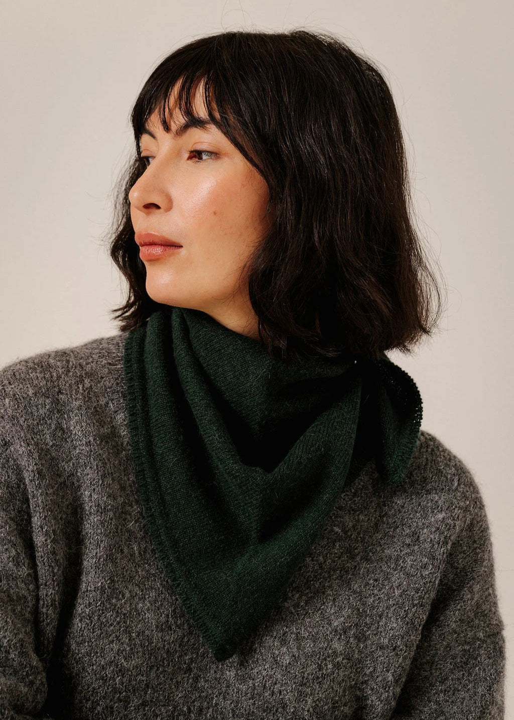 A woman with dark shoulder-length hair wearing a dark forest green triangular scarf and a textured gray knit sweater, photographed against a plain light beige background.