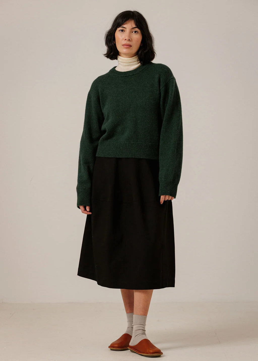 A model stands against a plain light beige studio background wearing a dark green knit sweater over a cream turtleneck, a black midi skirt, light gray socks, and brown slip-on shoes.
