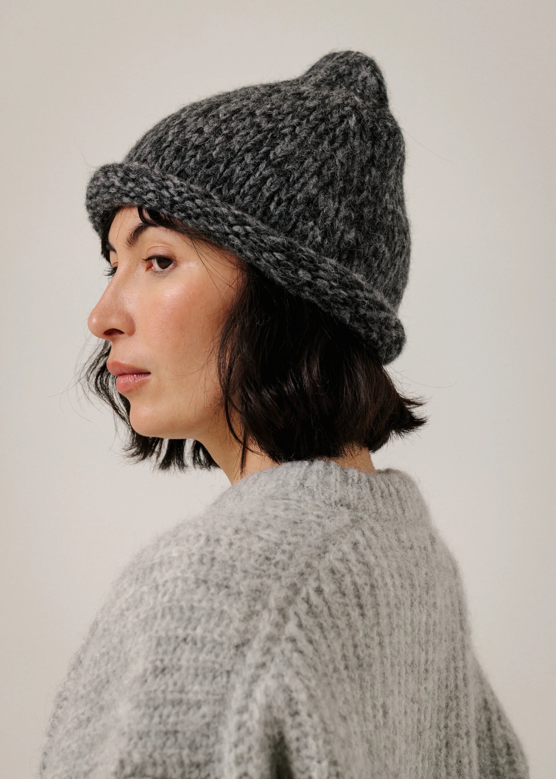 A woman shown in profile wearing a dark gray marled knit beanie and a light gray chunky knit sweater against a plain light background.