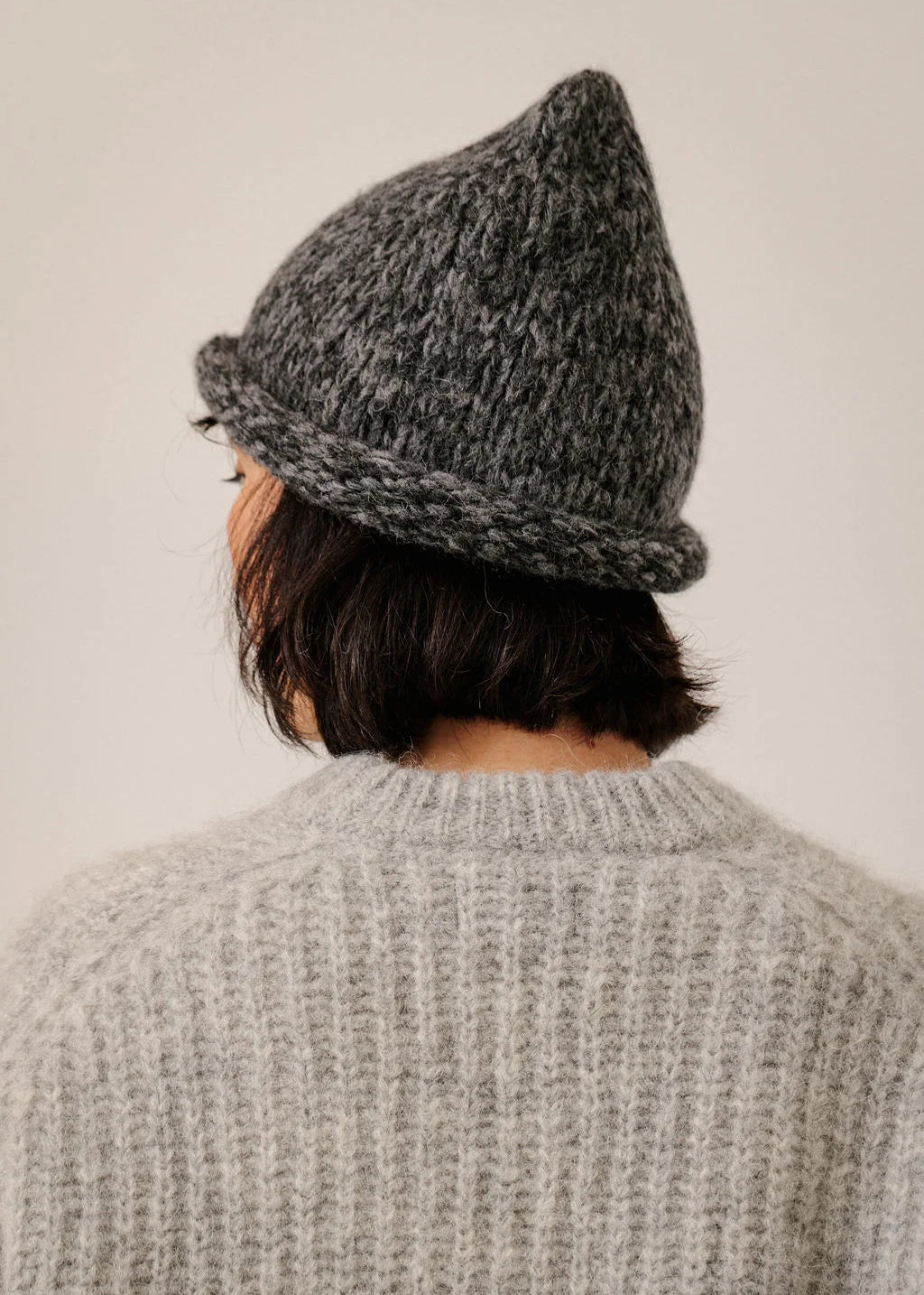A person shown from behind wearing a dark gray marled knit beanie and a light gray ribbed knit sweater against a plain light beige background.