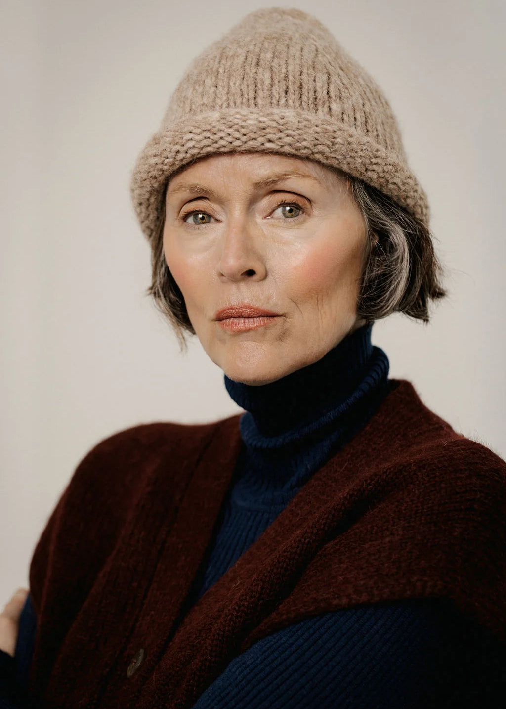 A close-up portrait of a woman wearing a beige chunky knit beanie, a dark navy blue turtleneck sweater, and a rust-brown knit cardigan against a plain light gray background.