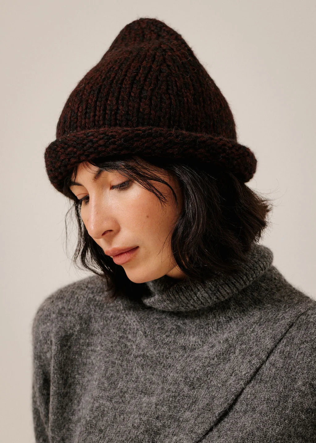 A woman wearing a dark brown chunky knit beanie and a gray turtleneck sweater, photographed against a plain light beige background.