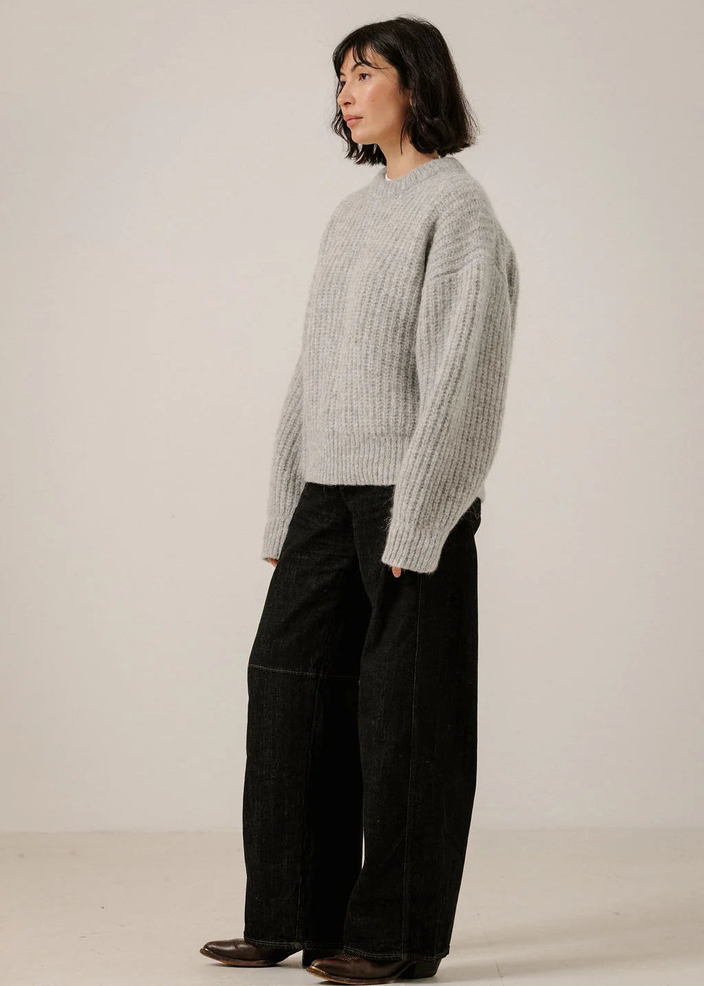 A model wearing a light gray ribbed knit sweater and black wide-leg pants, standing in profile against a plain off-white studio background.