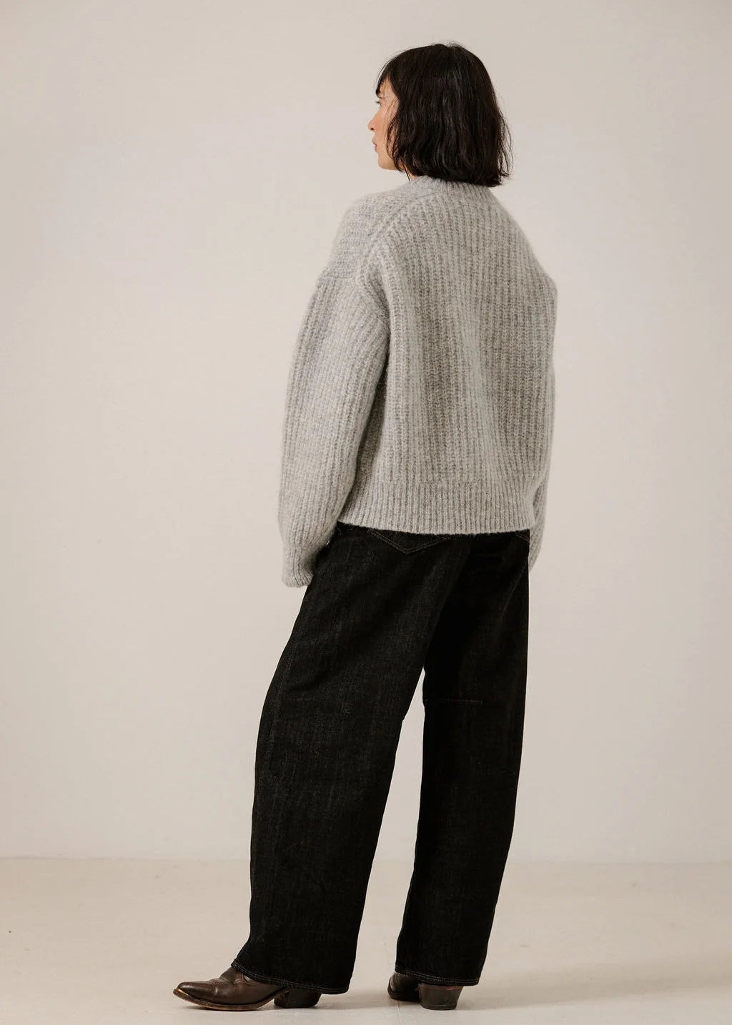 A model shown from the back wearing a light gray ribbed knit sweater and black wide-leg trousers against a plain off-white studio background.