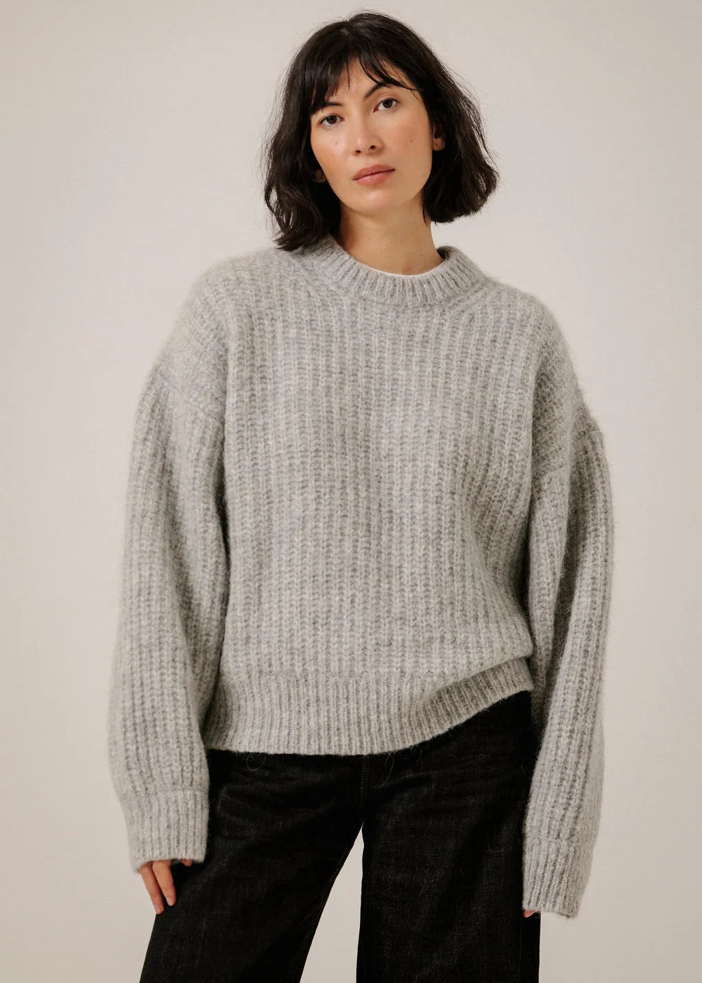 A woman wearing a light gray ribbed knit sweater and black pants, standing against a plain light gray studio background.