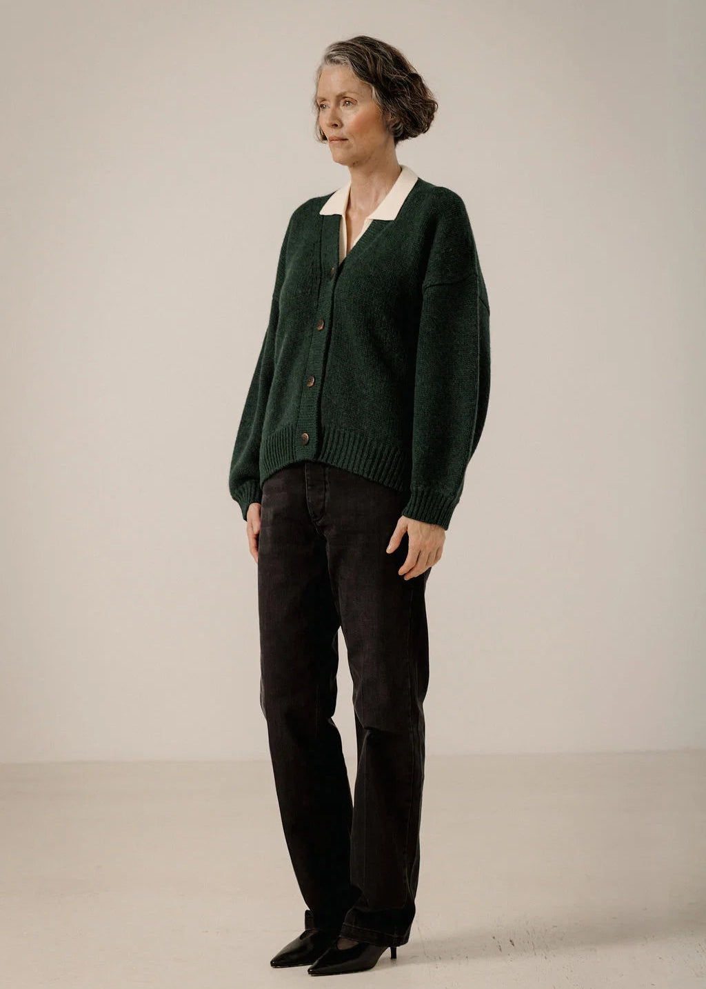 A woman stands in a studio wearing a dark green button-front cardigan over a cream collared top, paired with dark brown pants and black pointed shoes.