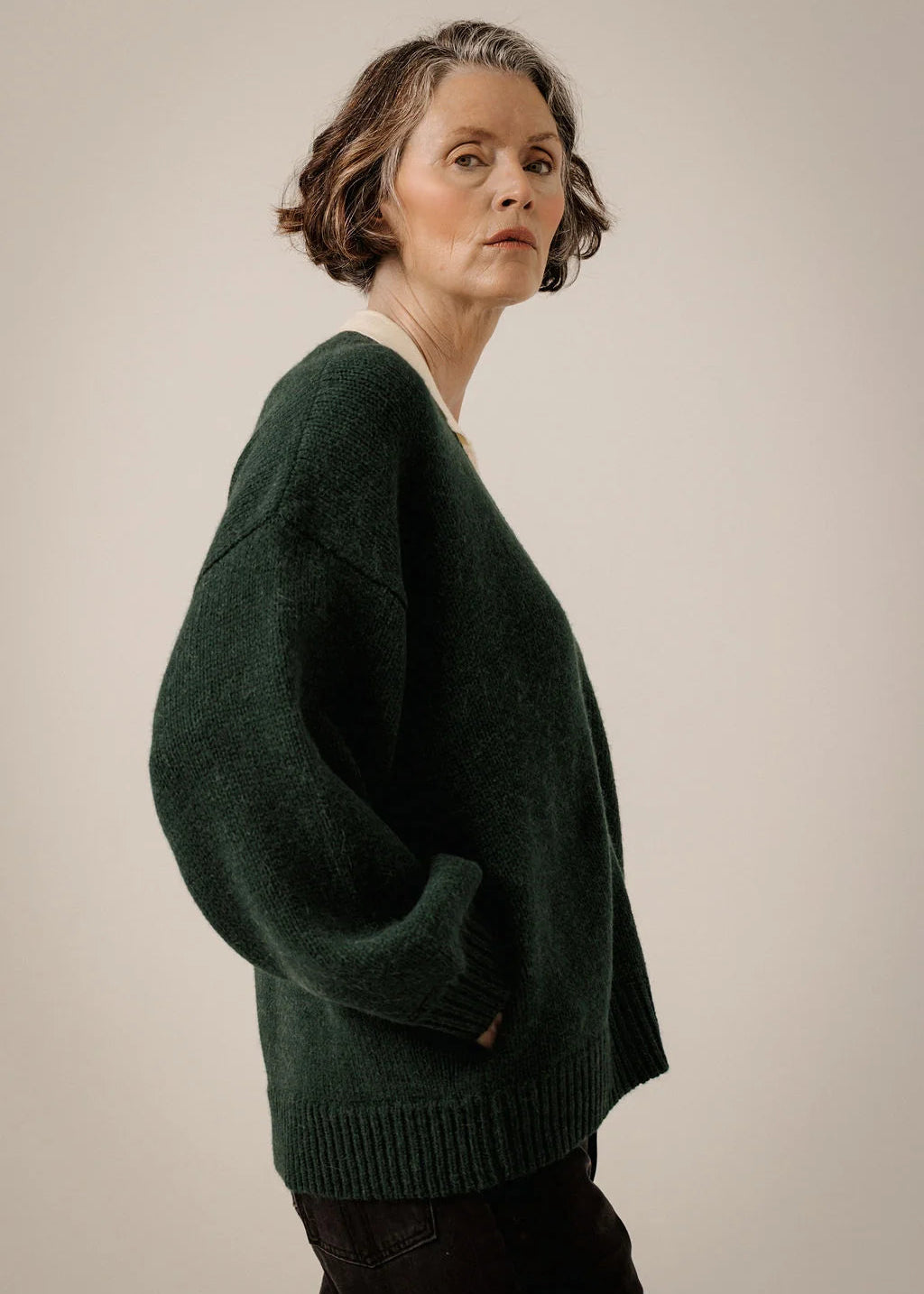 A woman wearing an oversized dark green knit sweater, shown from a side profile against a plain beige studio background.