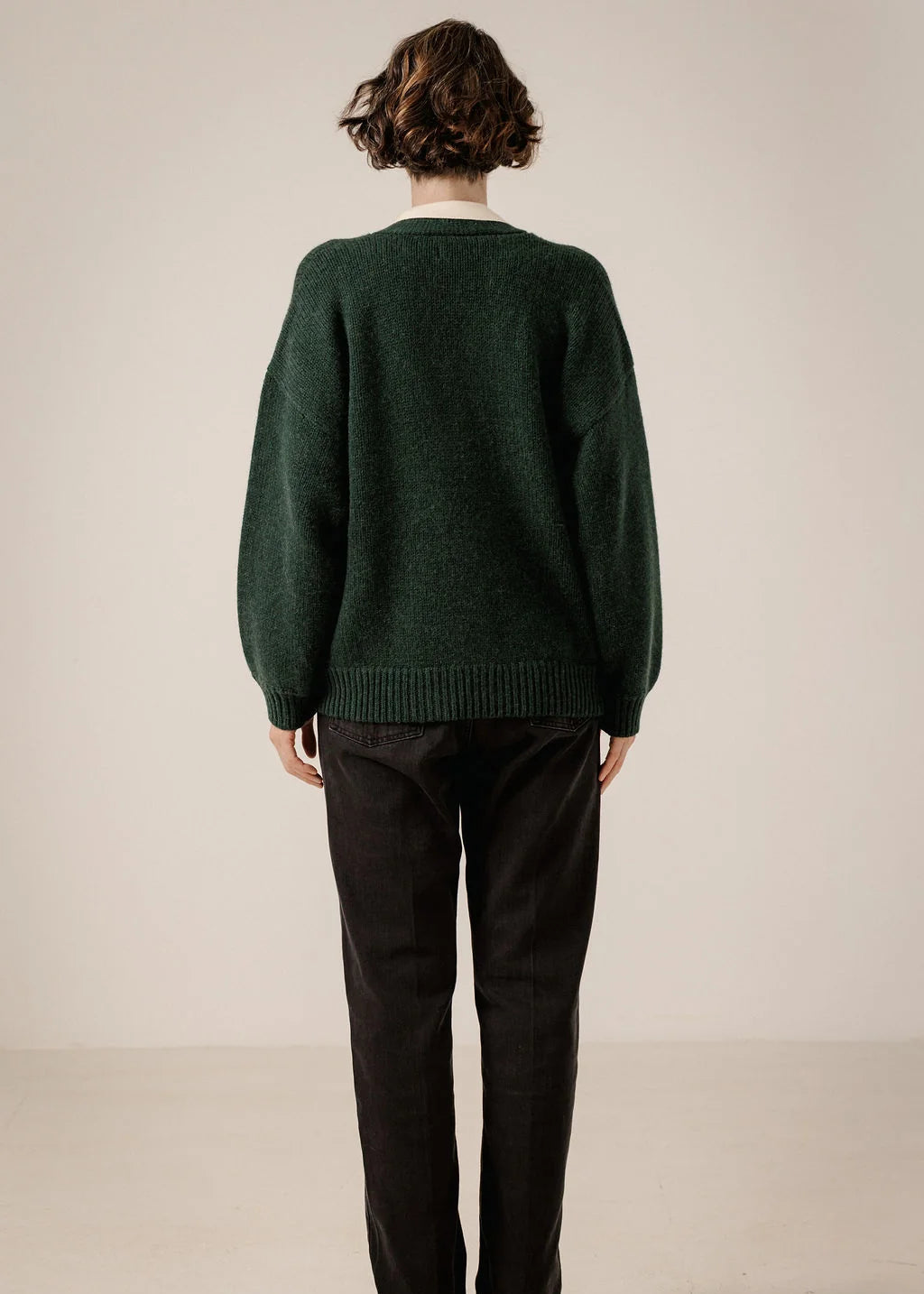 A person viewed from the back wearing an oversized dark green knit sweater and black pants against a plain light beige studio background.