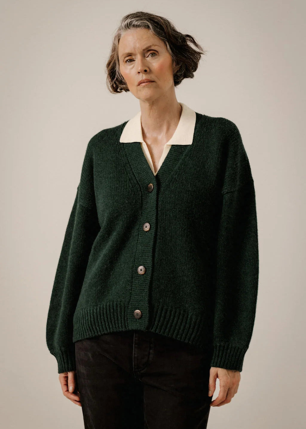 A woman wearing a dark green knit cardigan with a cream V-neck collar, paired with black pants, photographed against a plain light gray studio background.