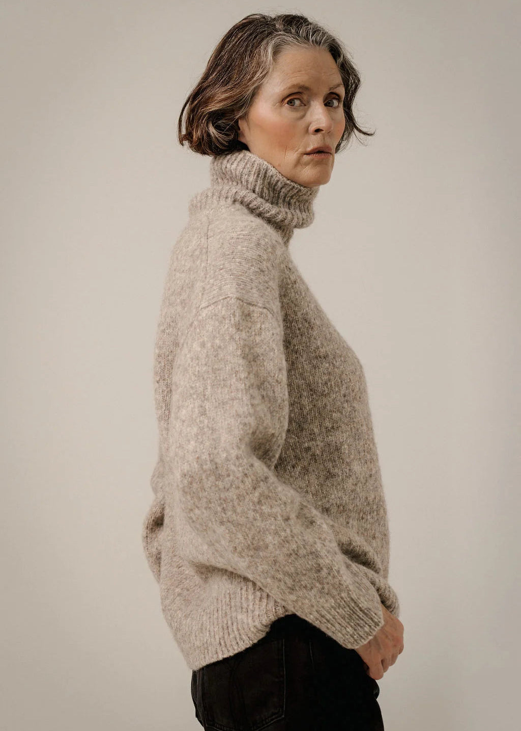 A woman wearing a chunky, textured oatmeal-beige turtleneck sweater, shown in a side profile against a plain, warm off-white studio background.