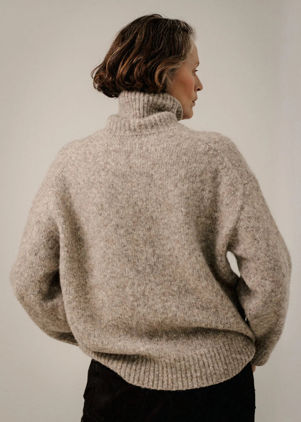 A woman shown from the back wearing an oversized, fuzzy turtleneck sweater in a heathered light taupe-beige color, paired with black pants, against a plain light gray studio background.